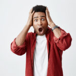 Portrait of young dark-skinned amazed man with afro hairstyle in white t-shirt under red shirt holding head with hands, looking in camera with opened mouth and shocked expression.